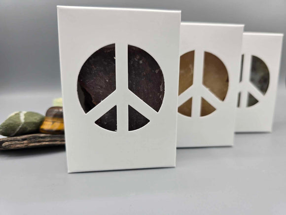 Three bars of plant based soaps in white soap boxes with a peace sign cut out. Behind the soap is driftwood and minerals.